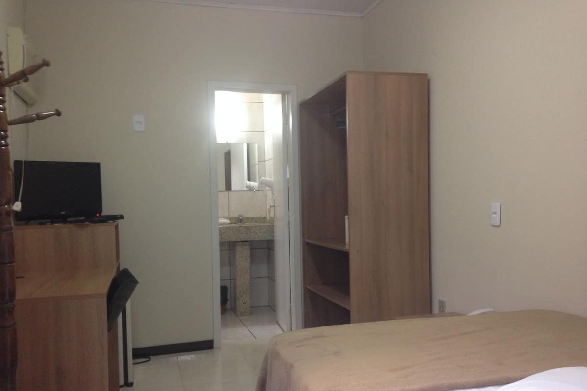 Quarto de hotel com uma cama, guarda-roupa de madeira, escrivaninha com televisão e banheiro visível através de uma porta aberta.