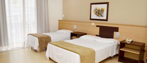 A hotel room with two single beds, a wooden headboard, a bedside table with a lamp, a framed picture on the wall, and a window with sheer curtains.