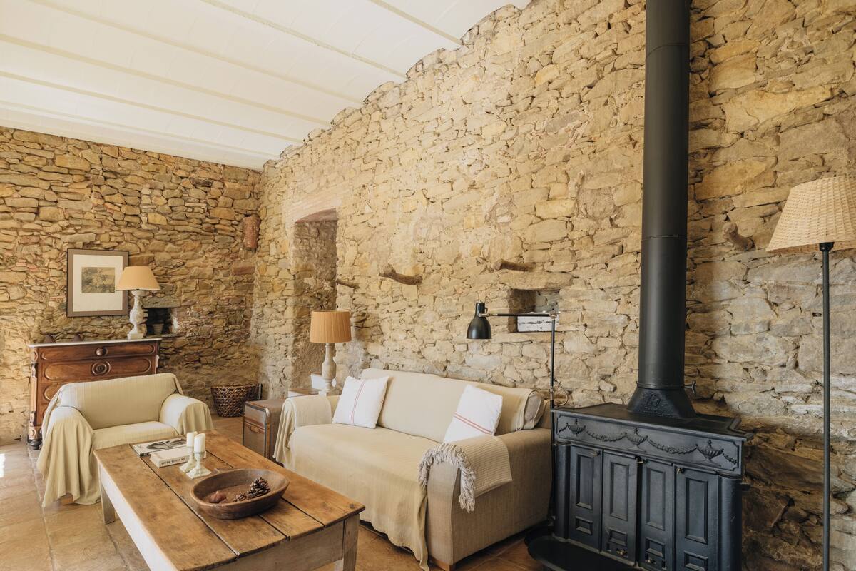 A cozy room with a stone wall, a wooden table, a sofa, and a black wood-burning stove.