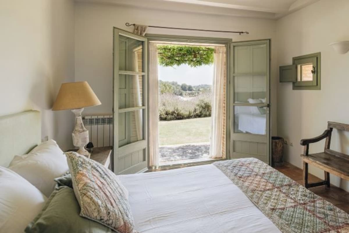 Un dormitorio con cama, lámpara, banqueta y ventana con vistas al exterior.
