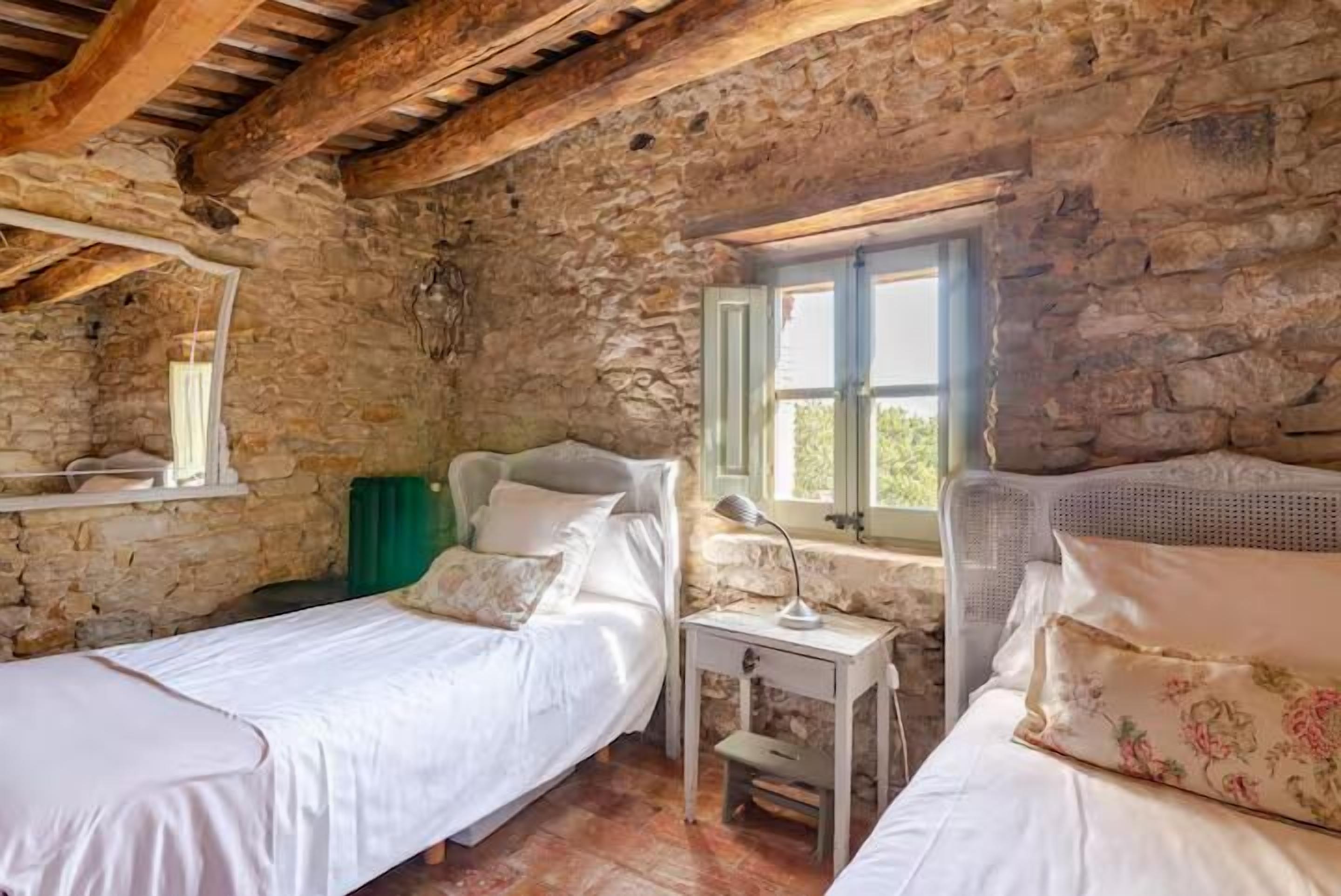Ein rustikales Zimmer mit Steinwänden, Holzbalken, zwei Betten mit weißer Bettwäsche, einem kleinen Tisch mit Lampe und einem Fenster mit grünen Fensterläden.