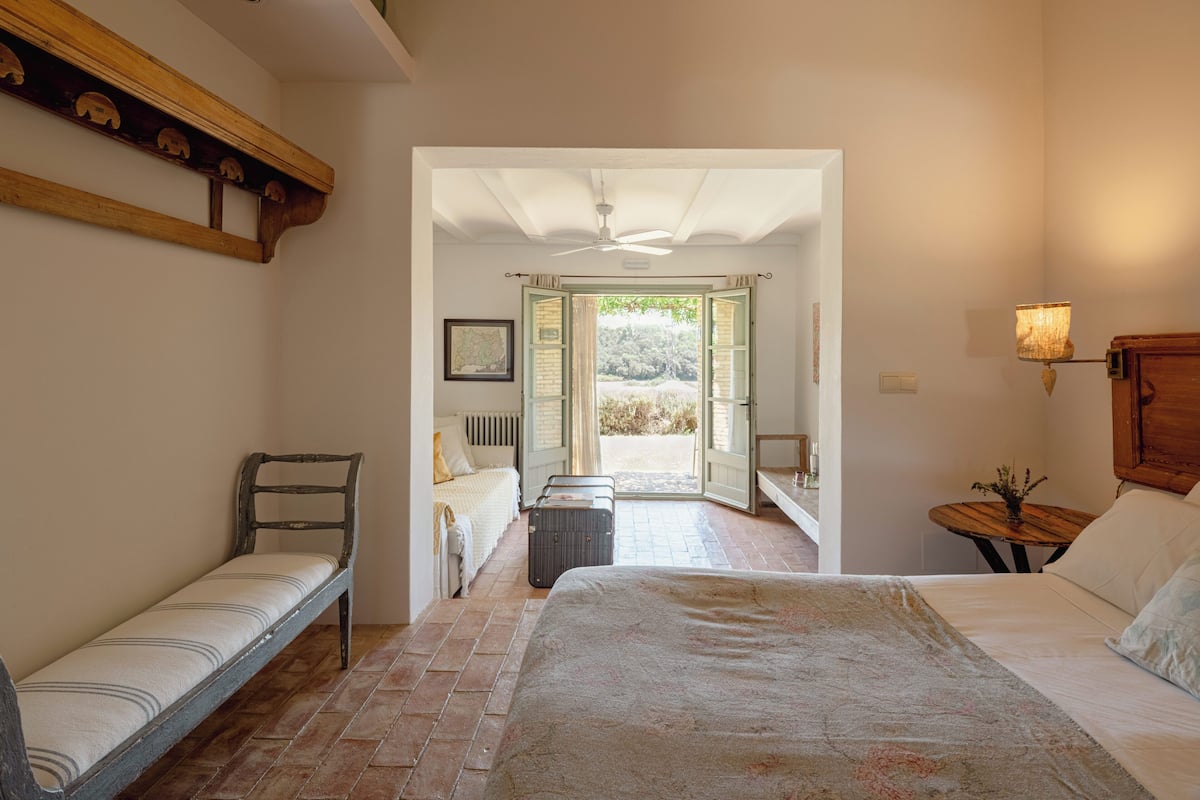 A bedroom with a large bed, a bench, a small table, and a view of the outside.