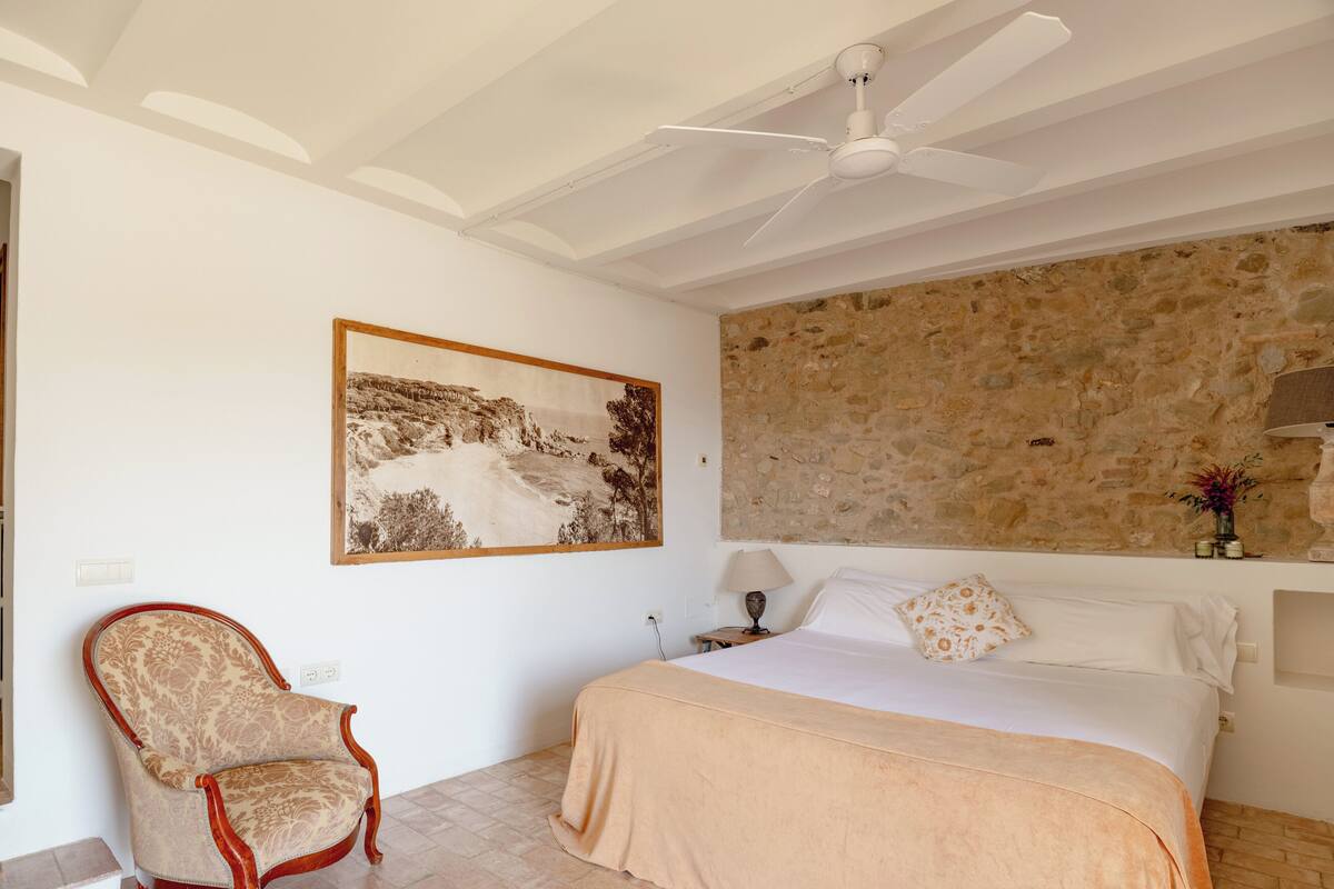 A bedroom with a bed, a chair, a ceiling fan, a stone wall, and a framed picture on the wall.