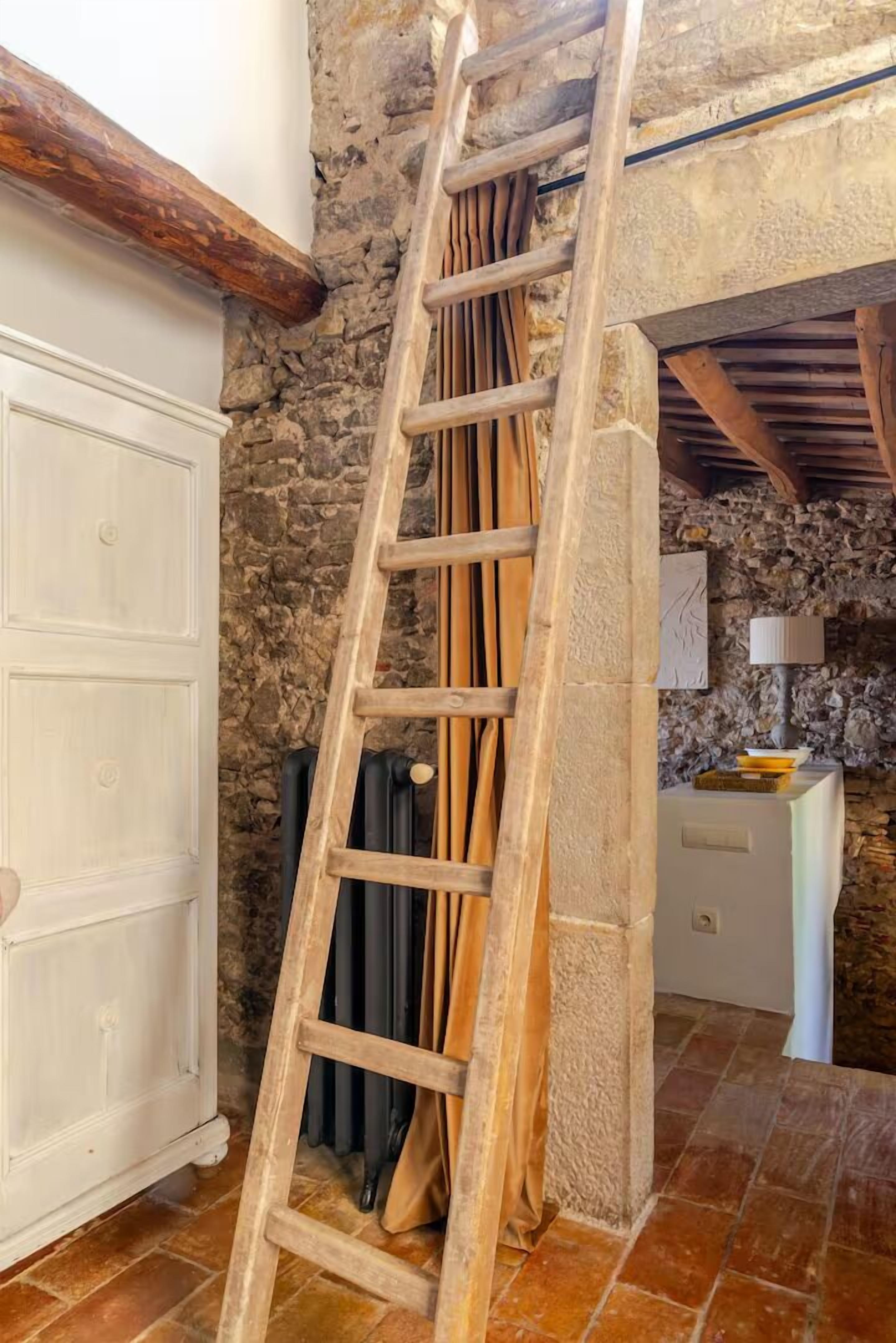 Une échelle en bois appuyée contre un mur de pierre, dans une pièce rustique où se trouvent une armoire blanche et une petite table avec une lampe.