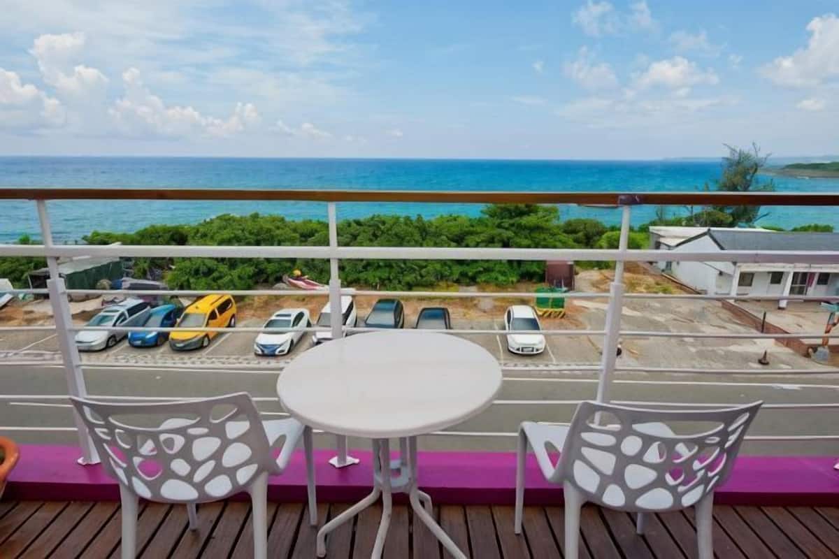 A balcony with a table and chairs, overlooking a parking area and the ocean.
