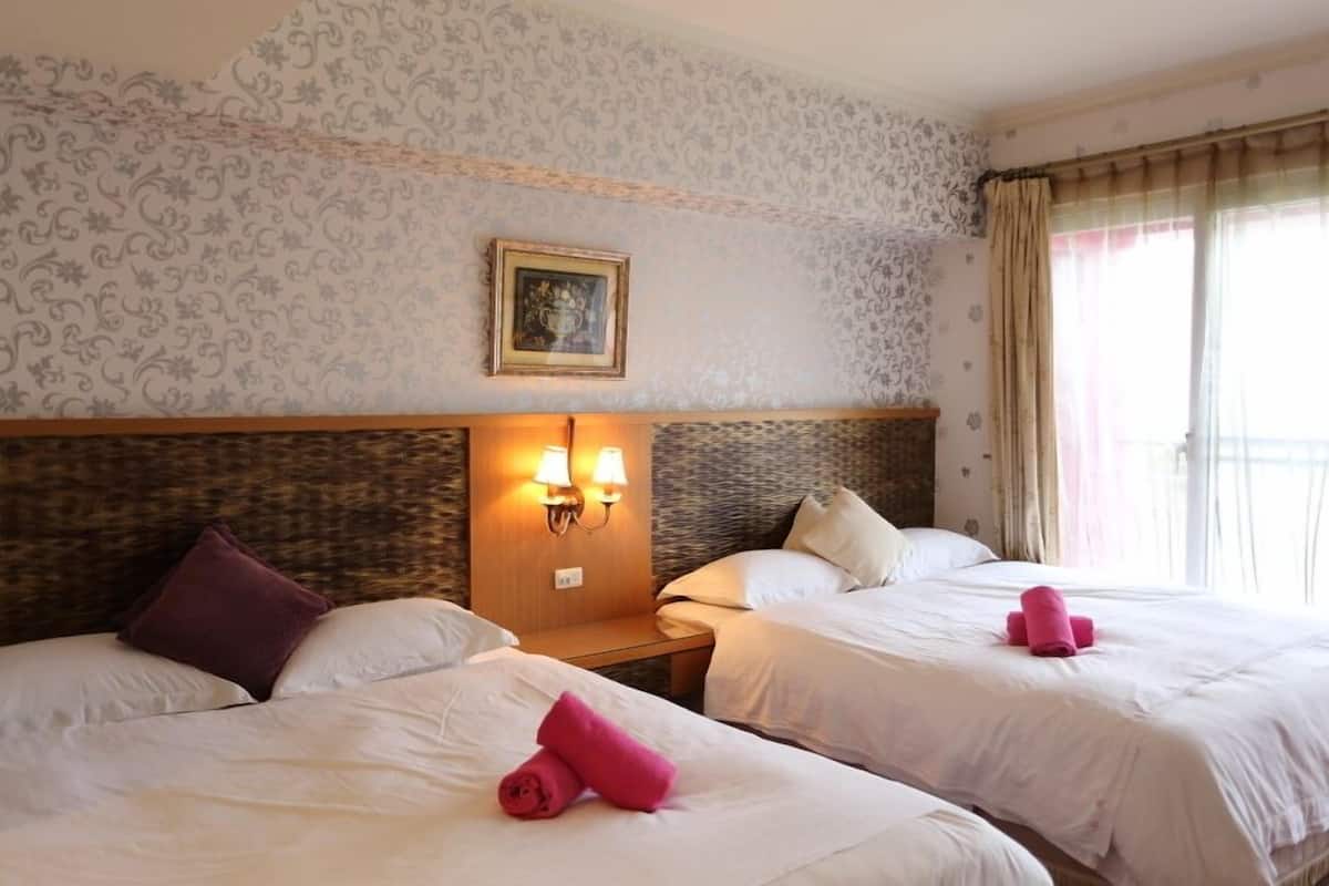 A hotel room with two beds, a headboard with wall sconces, a framed picture, and a window with curtains.