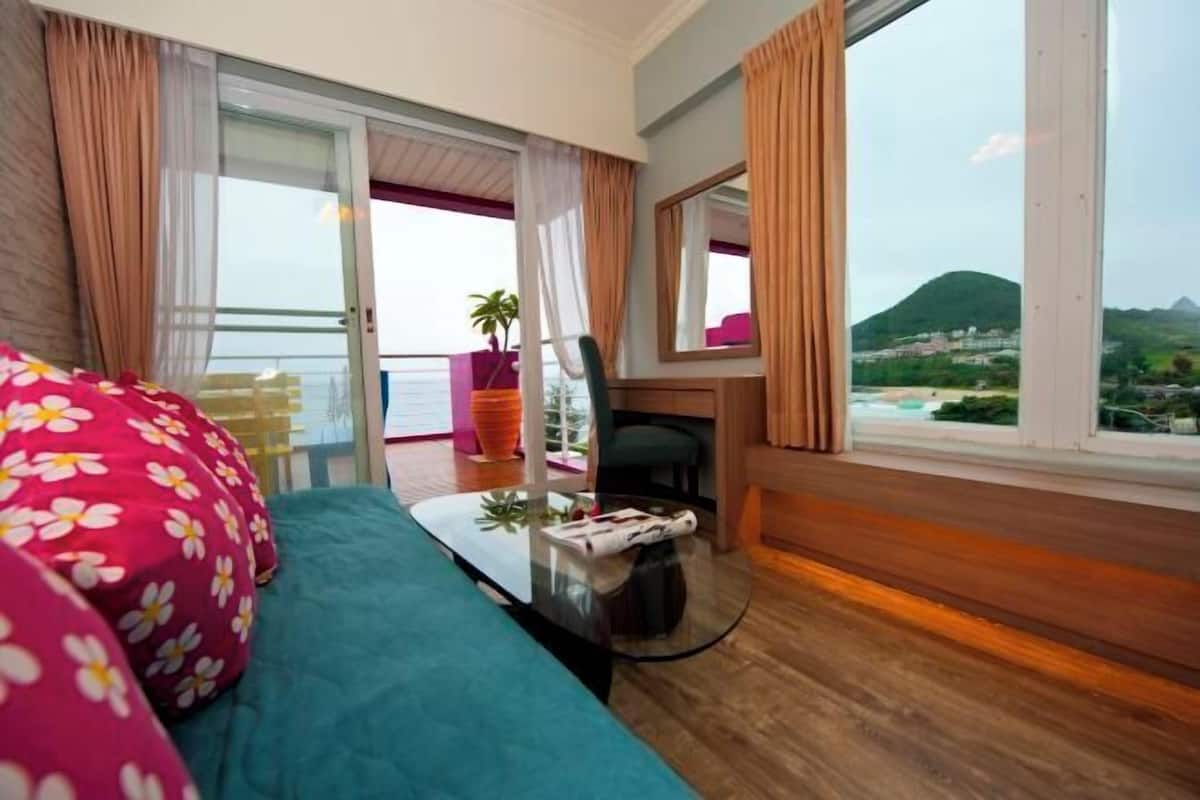 A hotel room with a bed, a small table, a chair, a window with a view of a mountain, and a balcony with a chair and table.