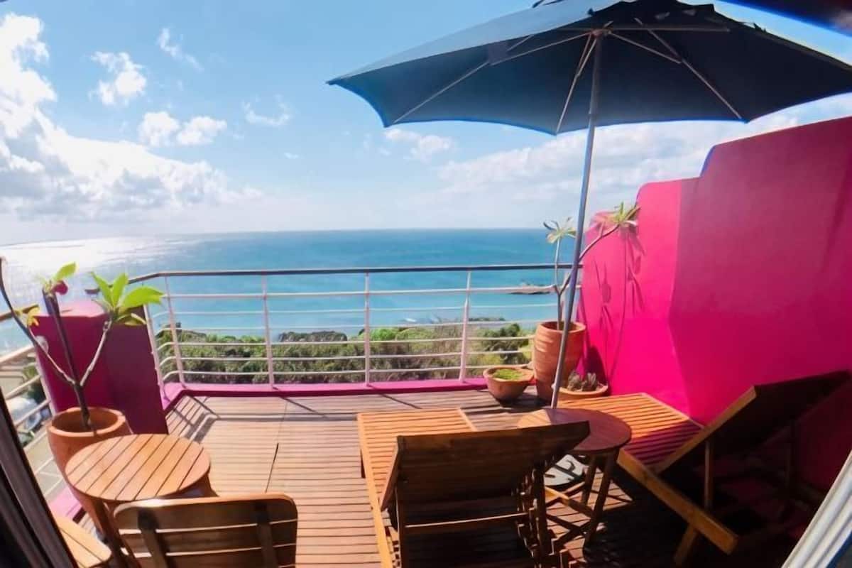 A balcony with a view of the ocean, wooden furniture, and a large umbrella.