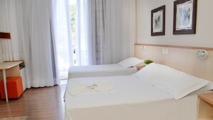 Habitación de hotel con dos camas, un escritorio de madera, un pequeño otoman naranja y vistas al verdor por la ventana.