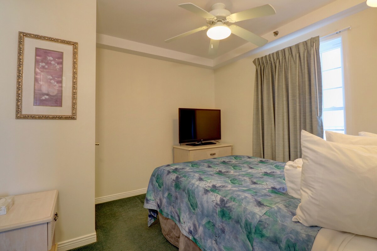 A hotel room with a bed, a television on a nightstand, a ceiling fan, and a window with curtains.