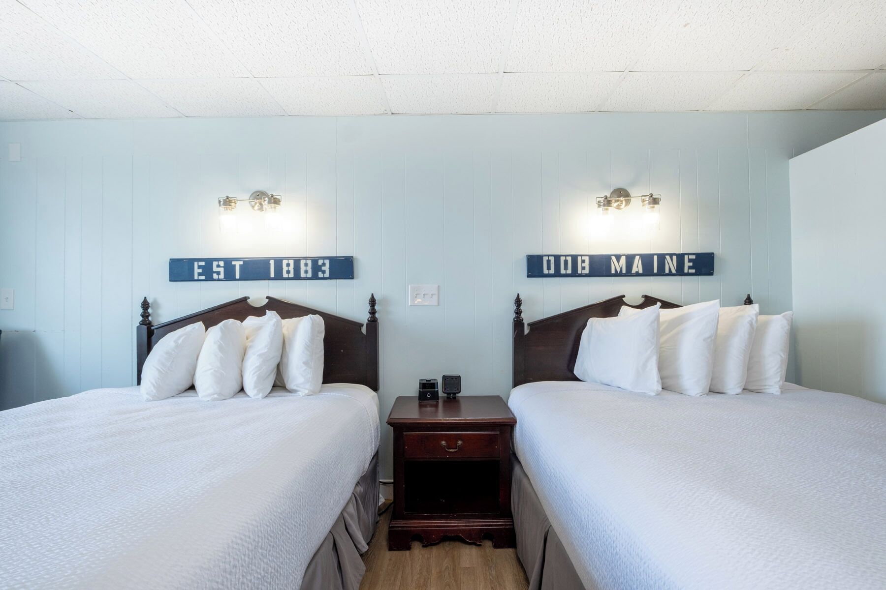 A room with two beds, each with a wooden headboard and white bedding. There are wall-mounted light fixtures above each bed.