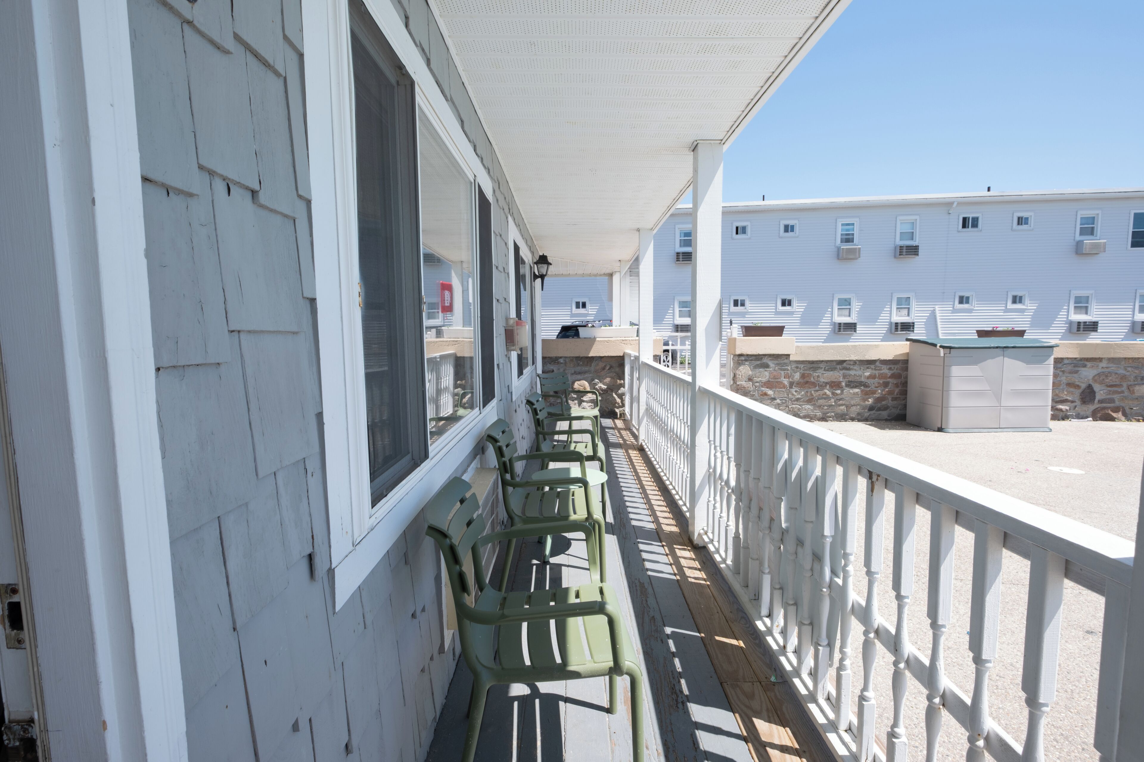 A covered balcony with rows of green chairs, white railings, and a view of adjacent buildings.