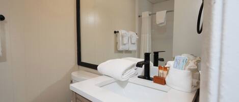 Un baño con un lavamanos blanco, un grifo negro y un espejo encima. Hay toallas colgadas en la pared y se ve un inodoro.