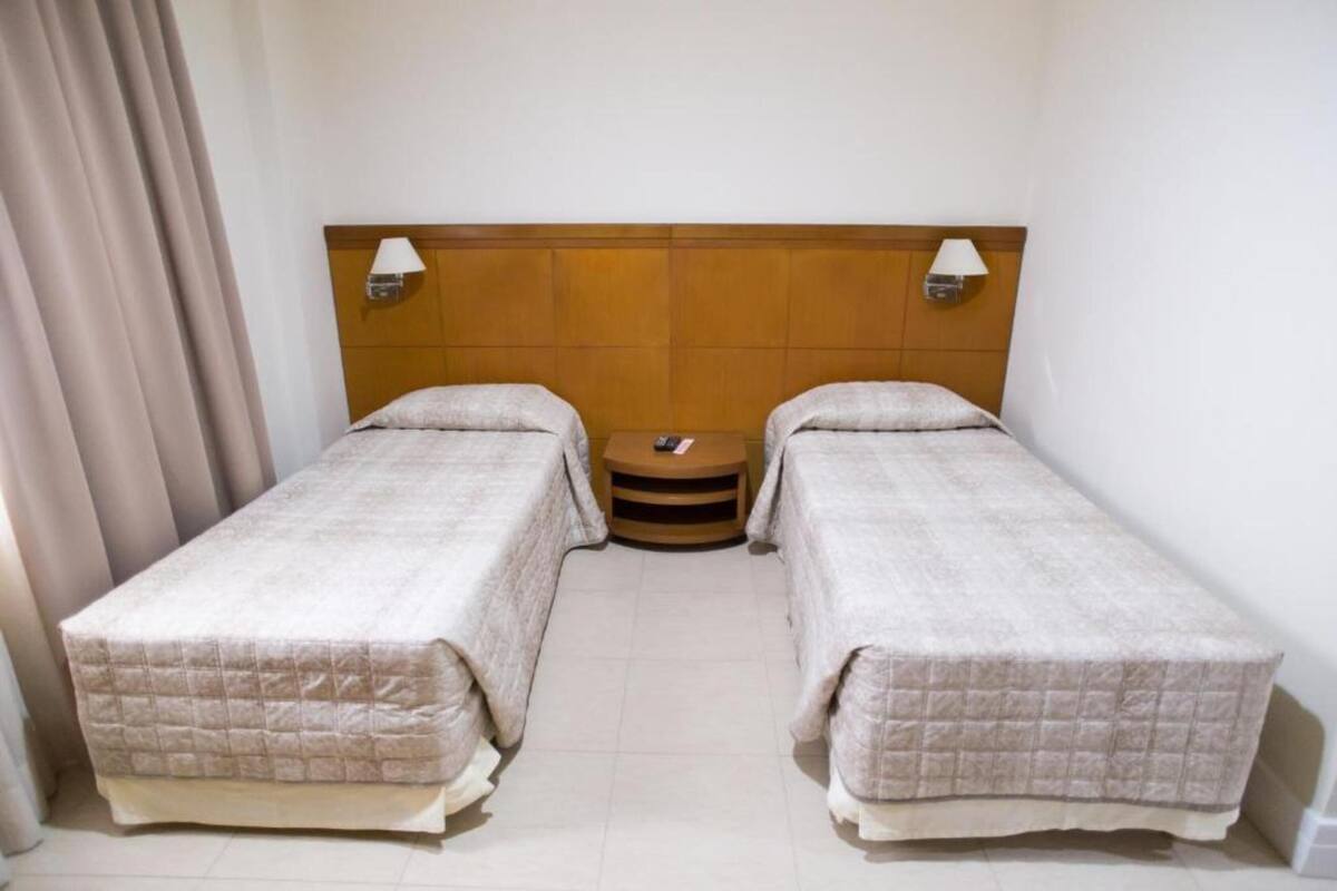 Een hotelkamer met twee enkele bedden, een houten hoofdbord, bedlampen, een klein rond tafeltje en een gordijn.