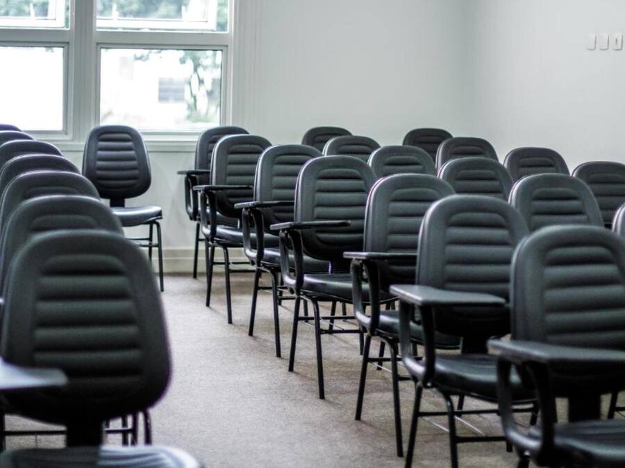Un salón de conferencias con filas de sillas negras y mesas, ventanas amplias con persianas y suelo alfombrado.