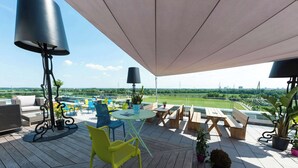 Un área de estar al aire libre con techo, mesas y sillas, vista a un campo verde y un horizonte urbano a lo lejos.