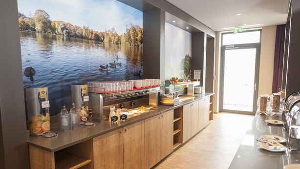 A modern hotel lobby with a large digital screen displaying a lake scene, a buffet counter with various food items, and a reception area with a door and signage.