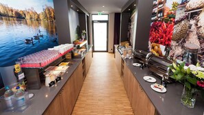 Zona de recepción de hotel con suelo de madera, una barra con diversas opciones de comida y una máquina de café, además de un mural que representa un lago con patos.
