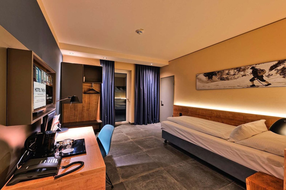 Habitación de hotel con cama, escritorio, teléfono, una silla y un cuadro en la pared.