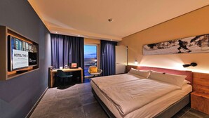 A hotel room with a bed, a desk, a chair, a small table, a nightstand, a wall-mounted lamp, and a window with curtains. - Best Western Hotel The K Munich Unterfoehring (Unterfoehring)
