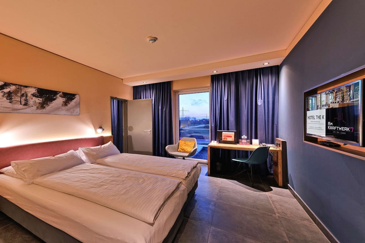 A hotel room with two beds, a desk, a chair, a TV, and a large window with curtains.