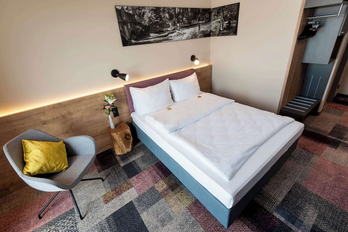 A modern hotel room with a large bed, a gray armchair with a yellow pillow, a small plant, and a wall-mounted artwork.