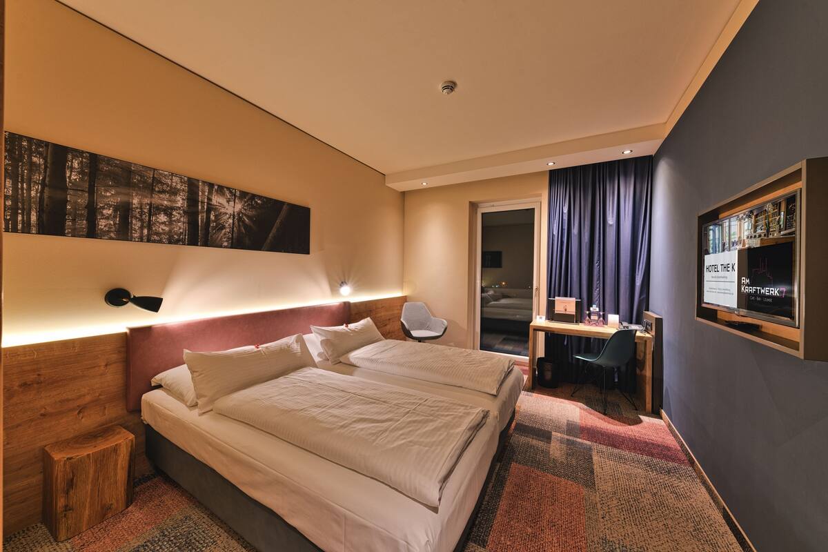 A hotel room with a large bed, a desk, a chair, a small stool, a wall-mounted TV, and a door leading to a balcony.