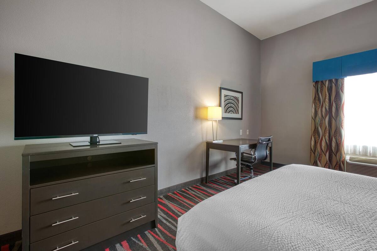 A hotel room with a bed, a desk with a chair, a television, a lamp, and a window with curtains.
