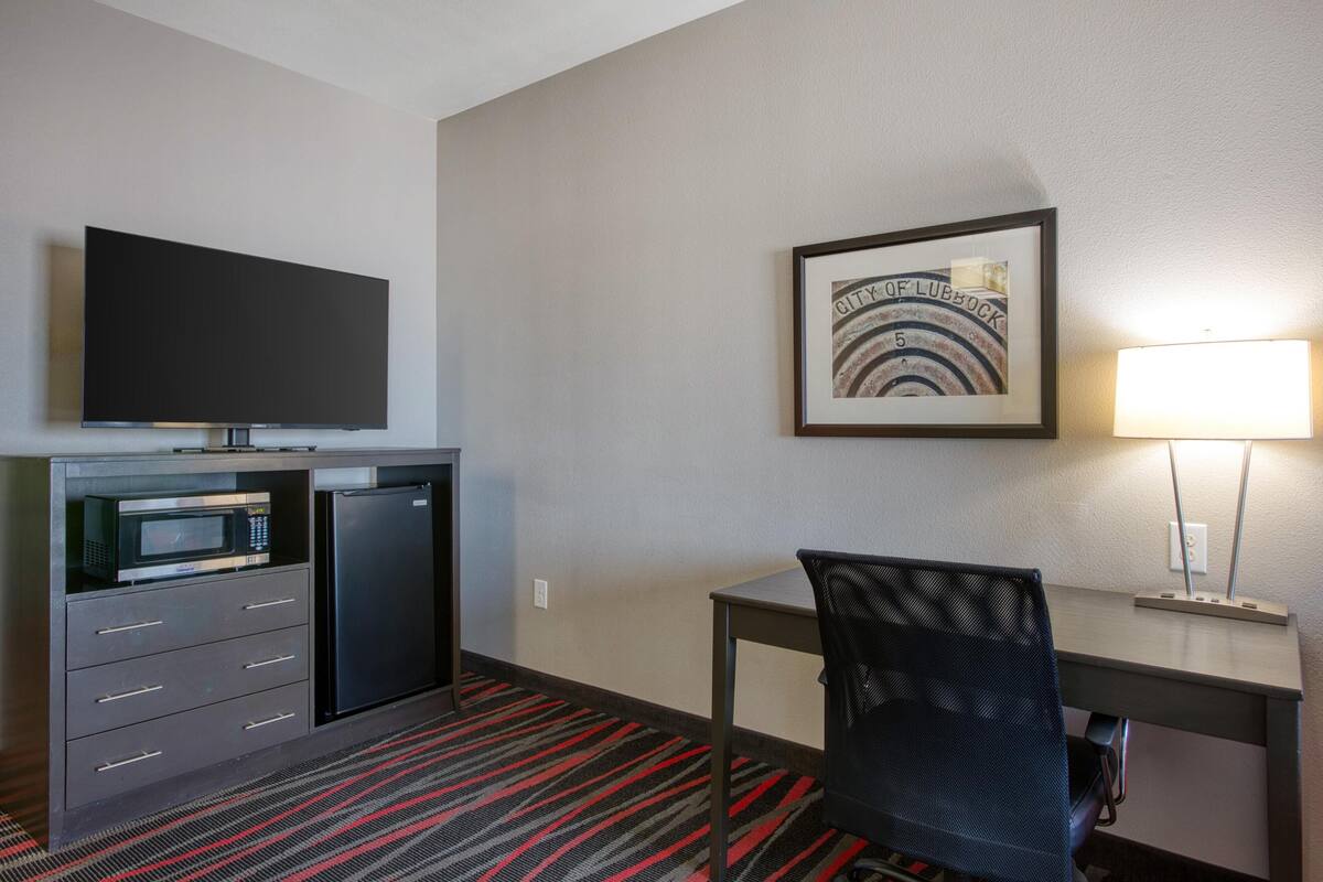 A hotel room with a flat-screen TV, a microwave, a refrigerator, a desk with a chair, a framed picture on the wall, and a lamp.
