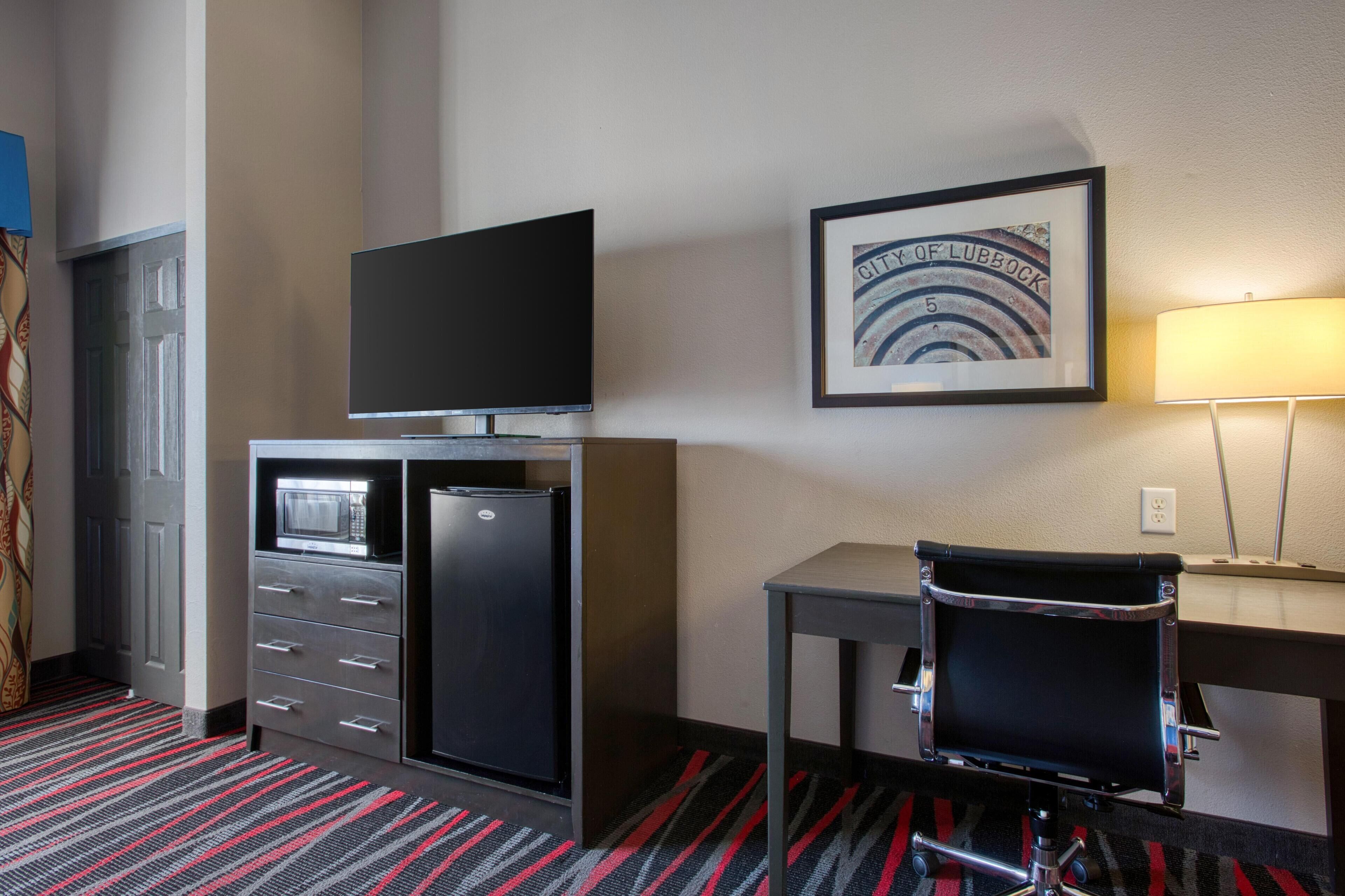 Habitación de hotel con televisión de pantalla plana en un mueble, un escritorio con una silla, un cuadro en la pared y una lámpara.
