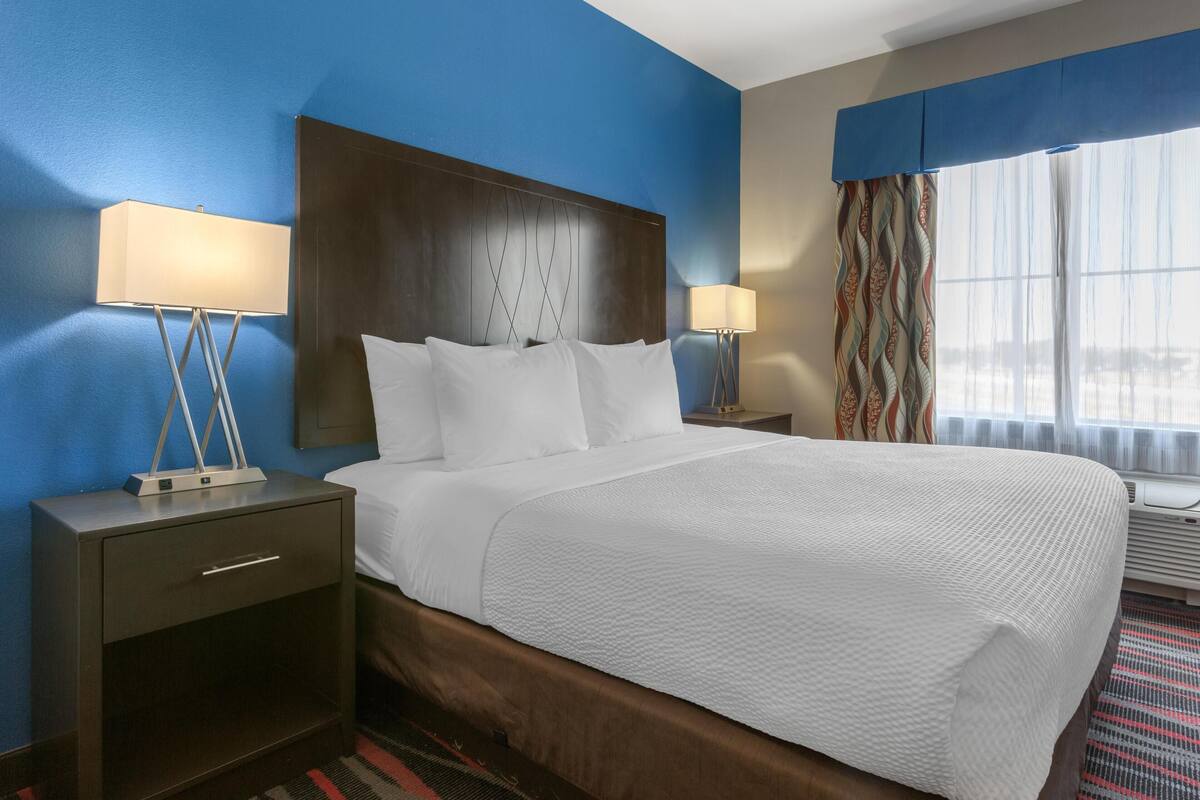 A hotel room with a large bed, a nightstand with a lamp, a dresser, and a window with curtains.