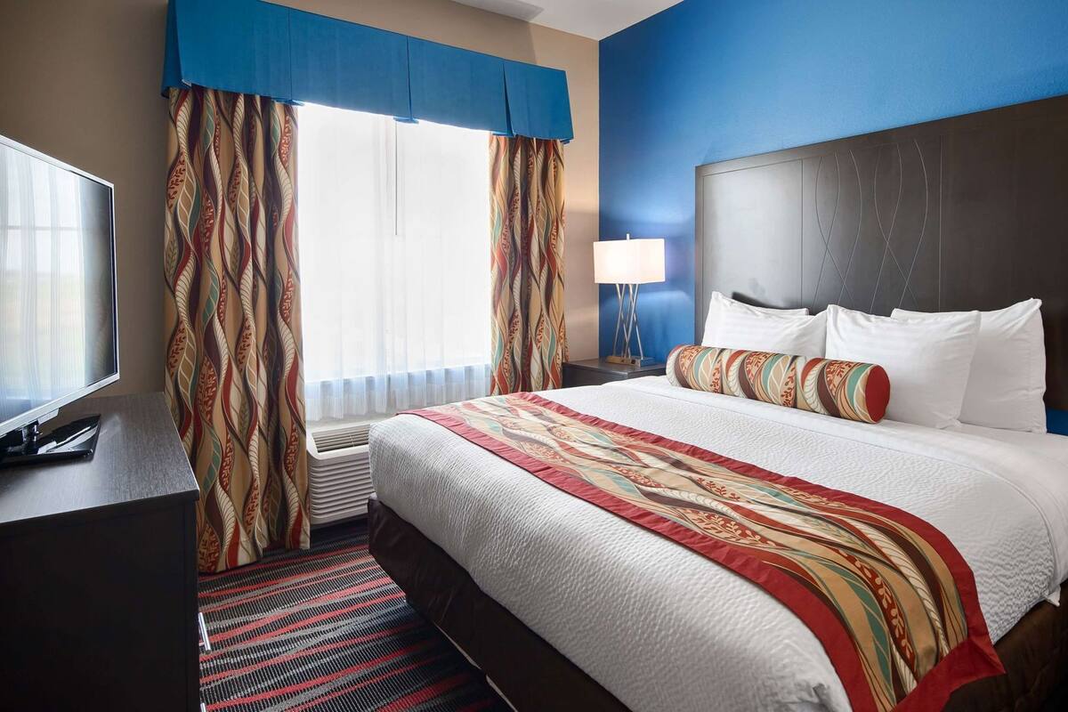 Ein Hotelzimmer mit einem großen Bett, einem Fernseher, einem Schreibtisch und einem Fenster mit Vorhängen.