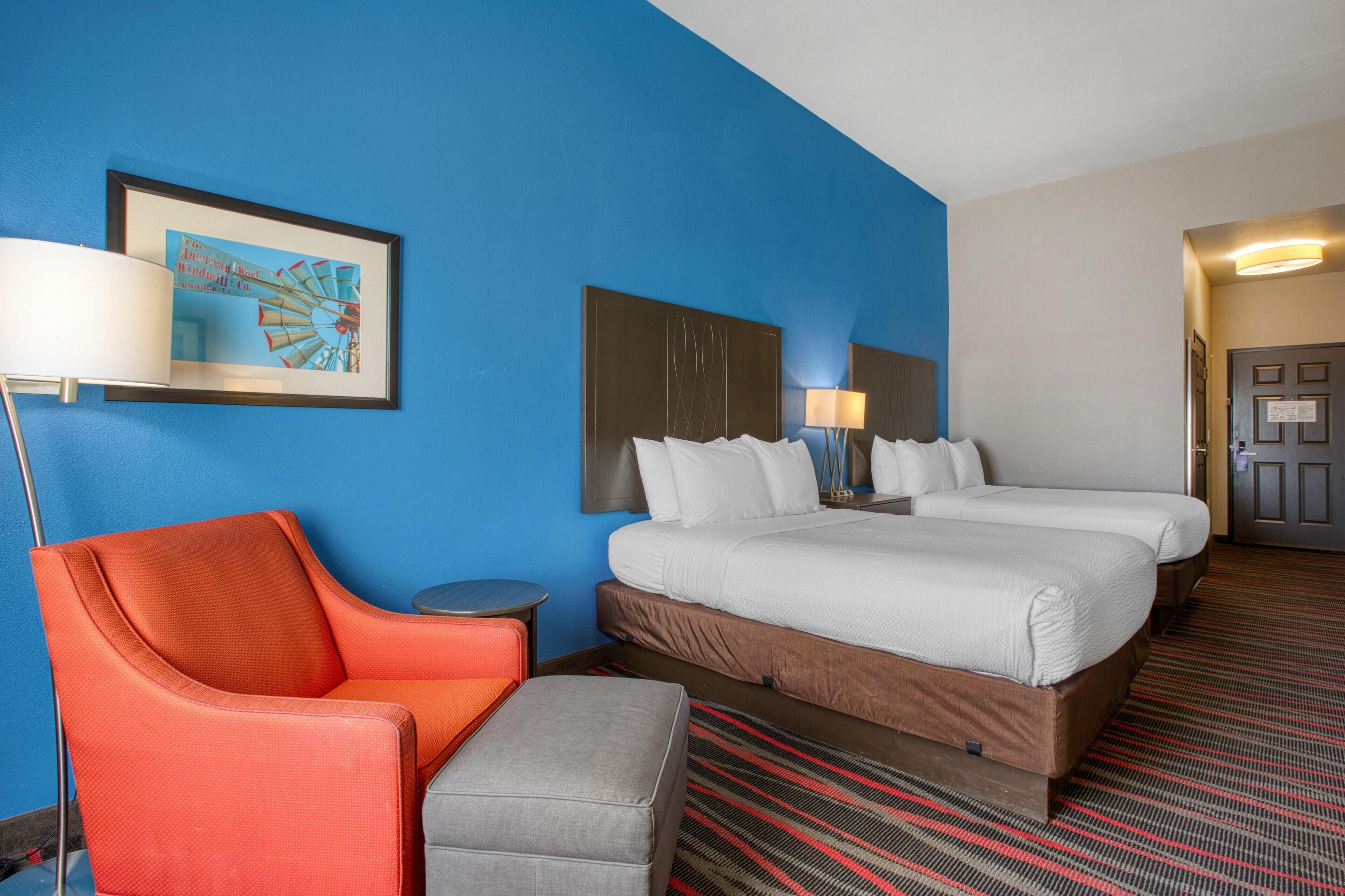 A hotel room with a large bed, two bedside tables with lamps, a red armchair, a grey ottoman, and a framed picture on the wall.
