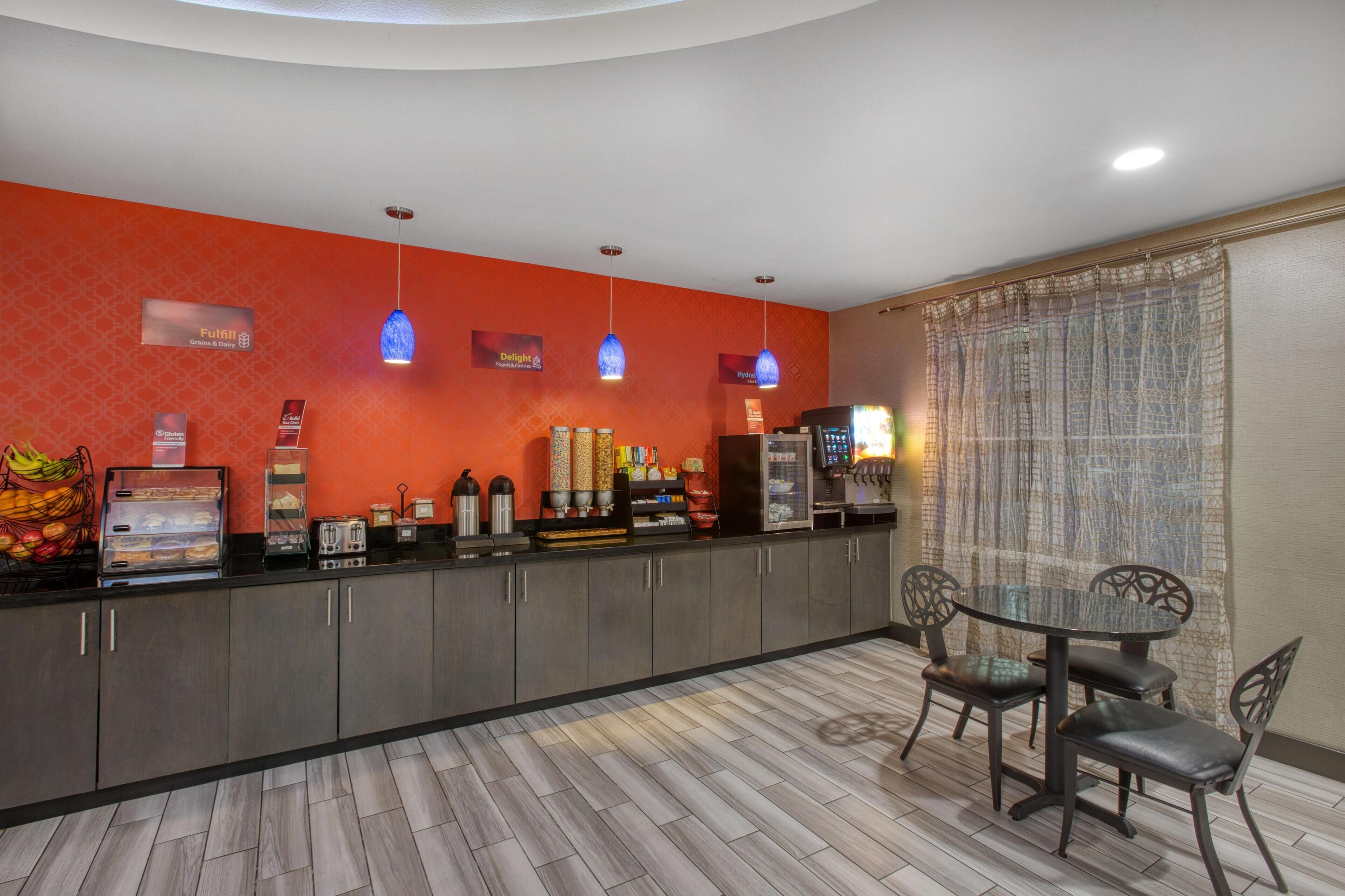 A hotel lobby with a red wall, a glass coffee station, and a seating area with a table and chairs.
