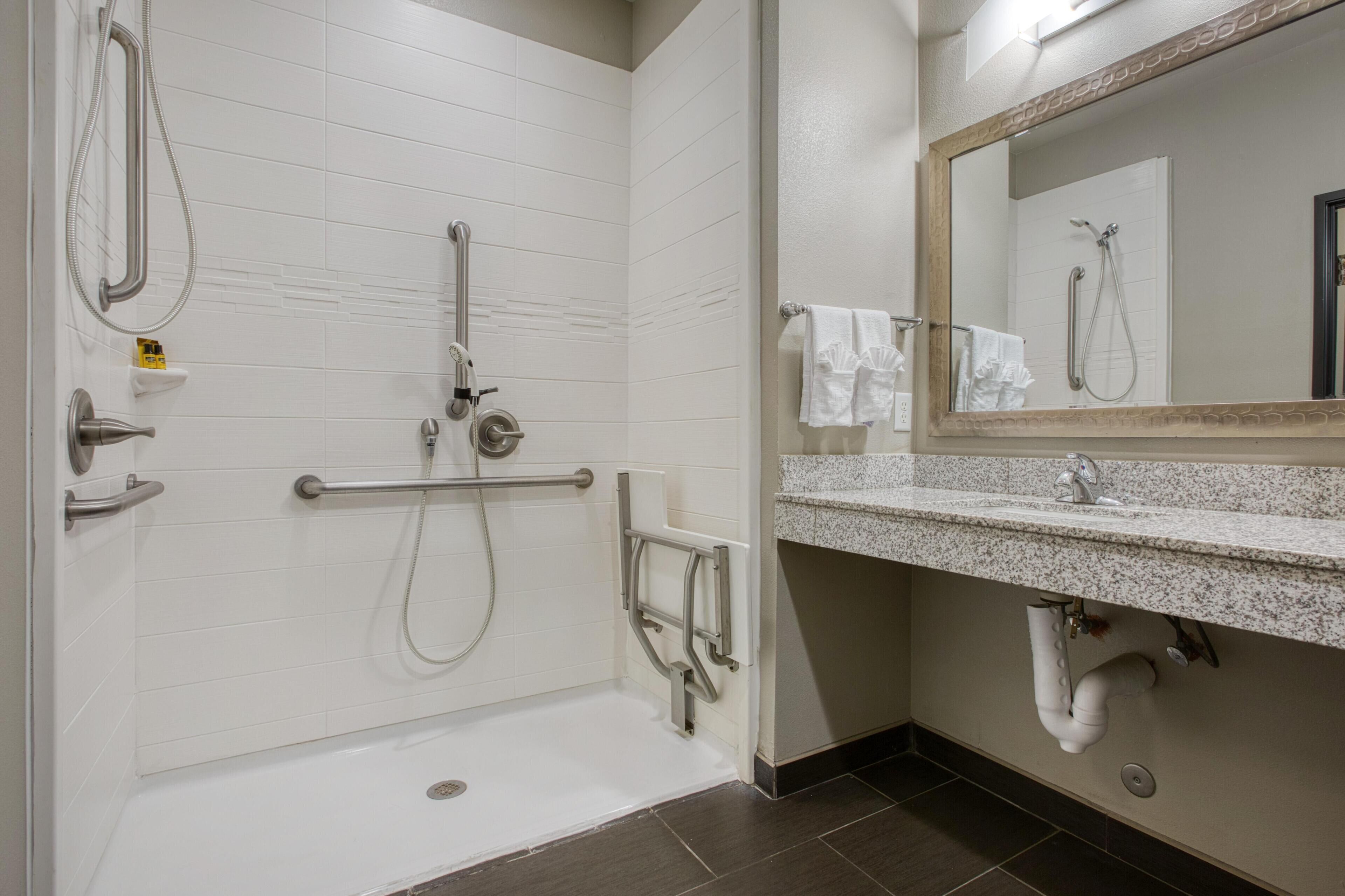 A modern bathroom with a walk-in shower, grab bars, a sink with a granite countertop, and a mirror above the sink.