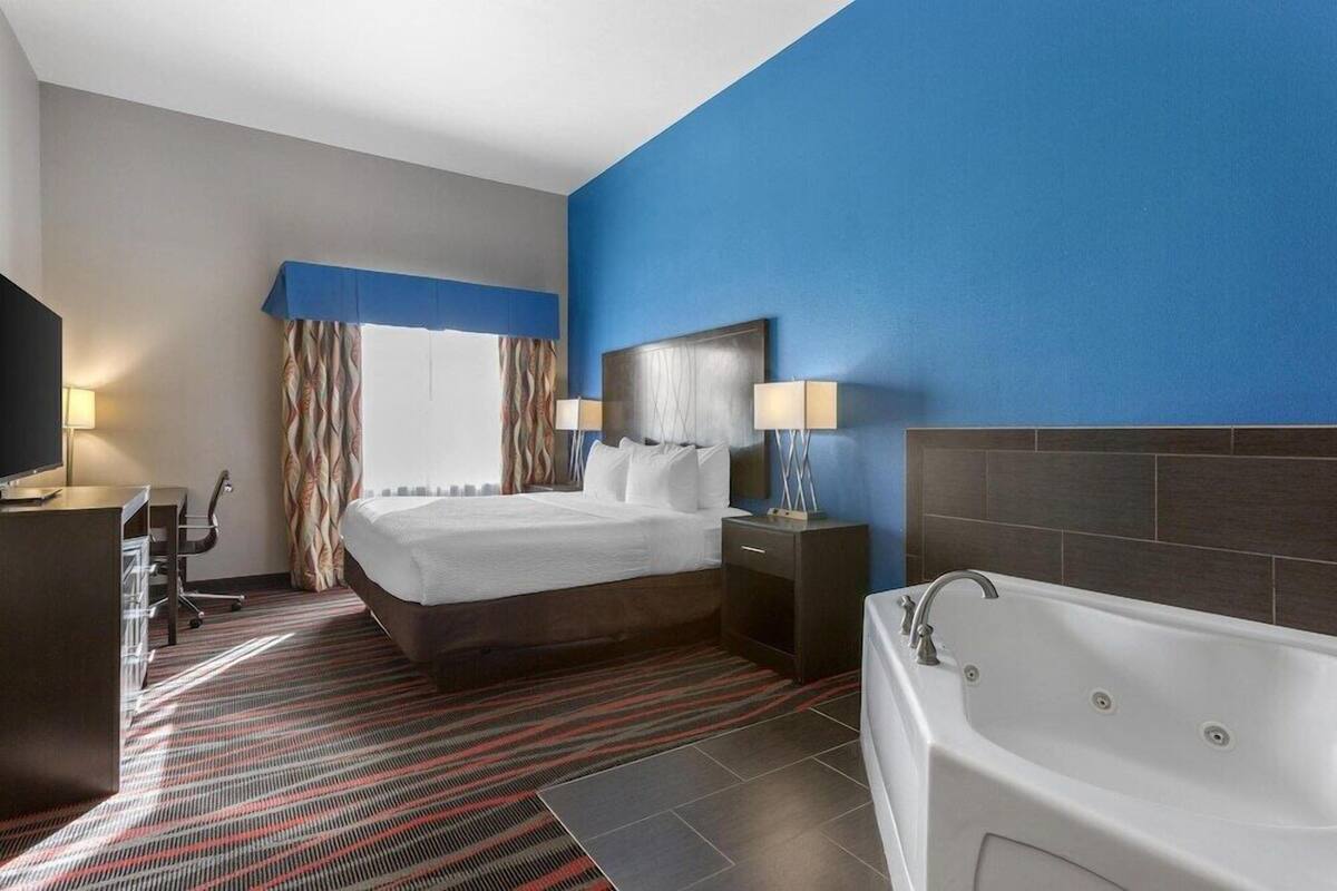 Ein Hotelzimmer mit Bett, Schreibtisch, Stuhl, Fernseher, einem Fenster mit Vorhängen sowie einem Badezimmer mit Badewanne und einer Wand in Blautönen.