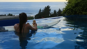 Eine Person im Pool, die eine Flasche hält, mit einem Surfbrett und Blick auf den Ozean.