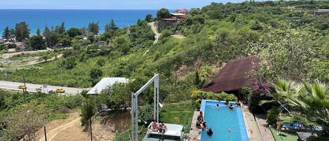 A pool area with a view of the ocean, surrounded by lush greenery and a hilly landscape.