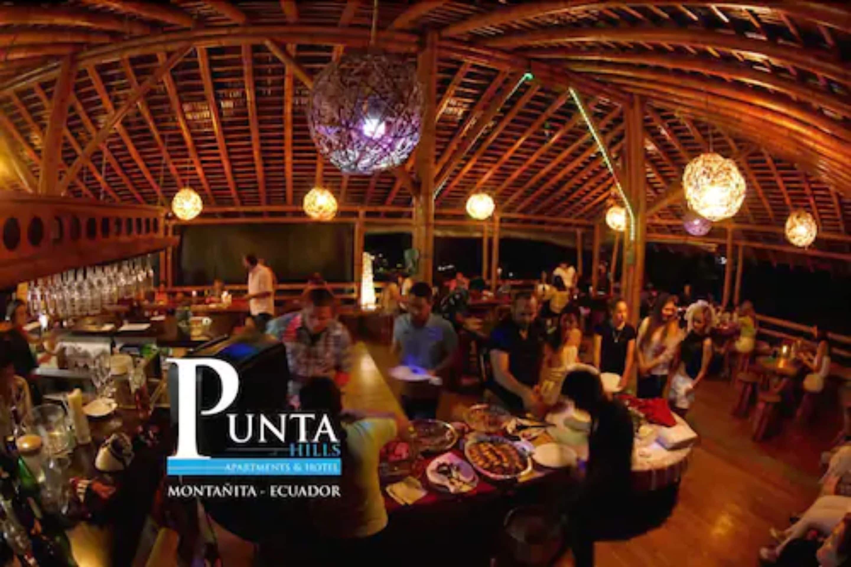 Un restaurante con techo de madera y luces esféricas colgantes. Las mesas están preparadas con platos y vasos.