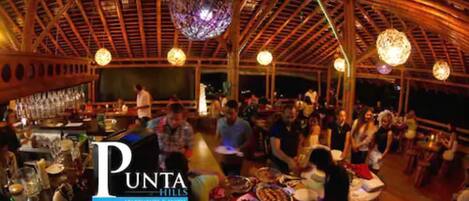 Un restaurante con techo de madera y luces esféricas colgantes. Las mesas están preparadas con platos y vasos.