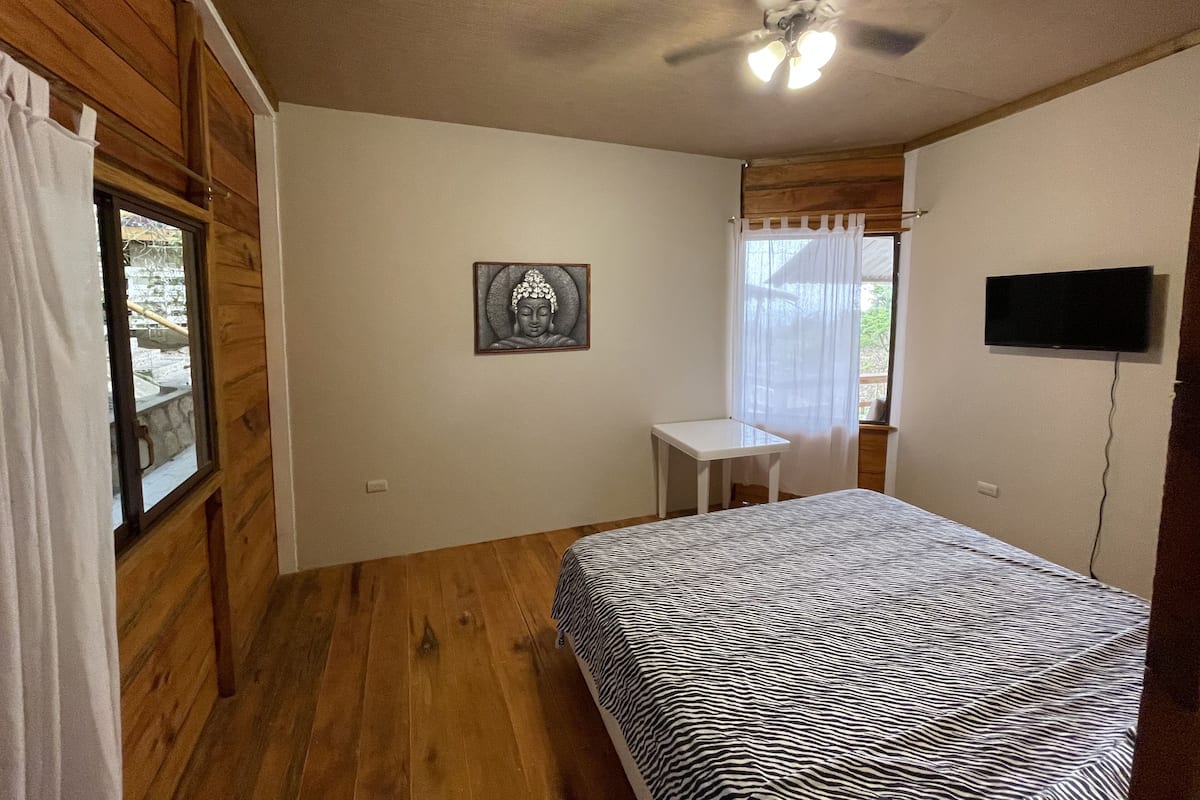 Une chambre avec un plancher en bois, un lit recouvert d’un couvre-lit à motifs zébrés, un ventilateur de plafond, une petite table blanche, un téléviseur fixé au mur et une fenêtre avec un rideau.