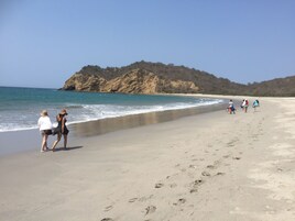 Una playa arenosa con huellas, una colina rocosa al fondo y un cielo azul despejado.