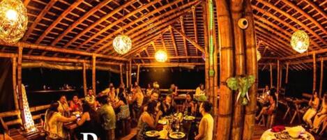 A wooden-roofed dining area with tables and chairs, bamboo pillars, and hanging light fixtures.