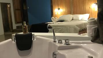 A bathroom with a large white bathtub, a wooden wall, and a bed with white bedding in the background.