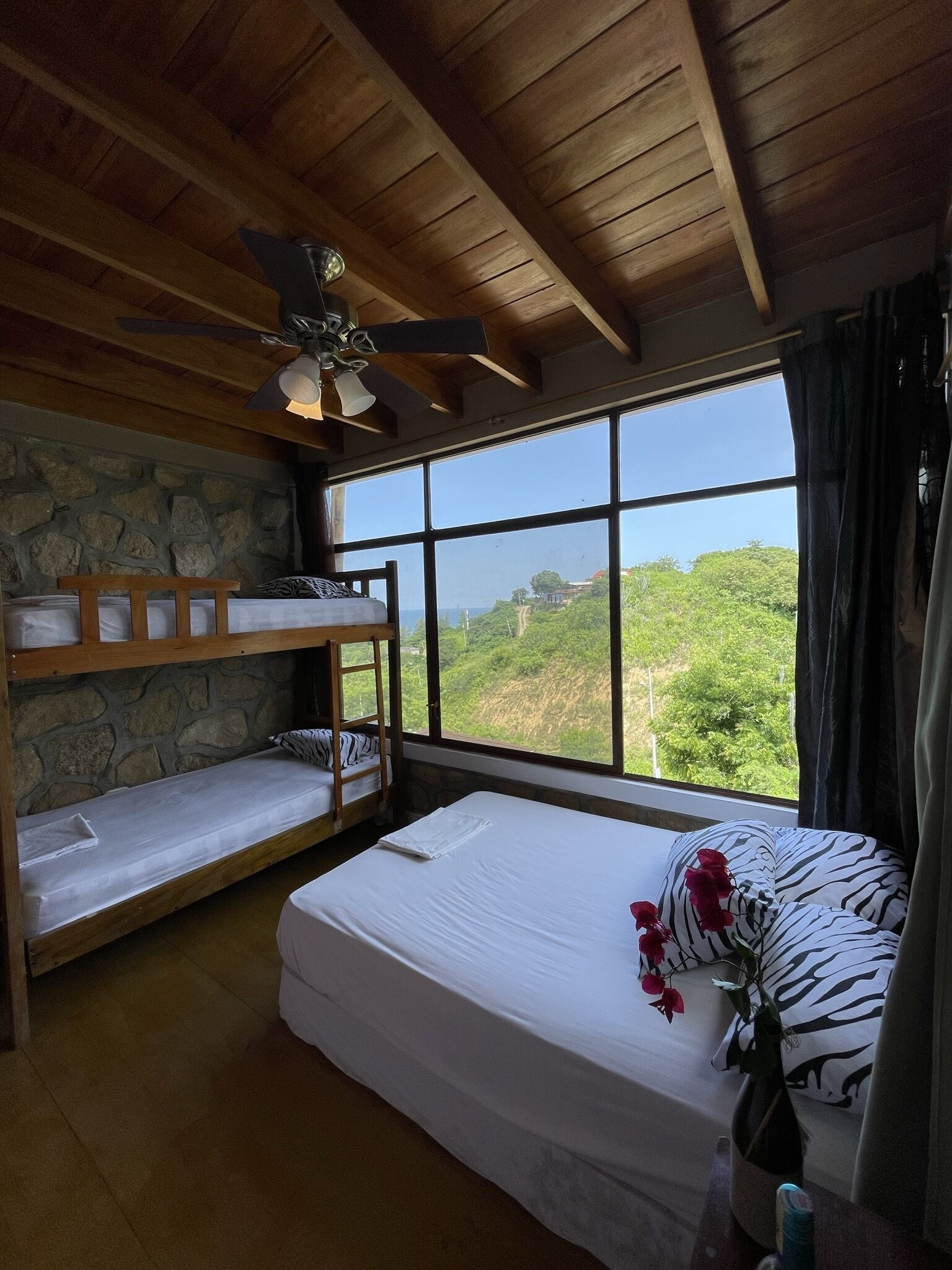A room with bunk beds, a ceiling fan, a large window with a view of greenery, and a bed with a flower arrangement.