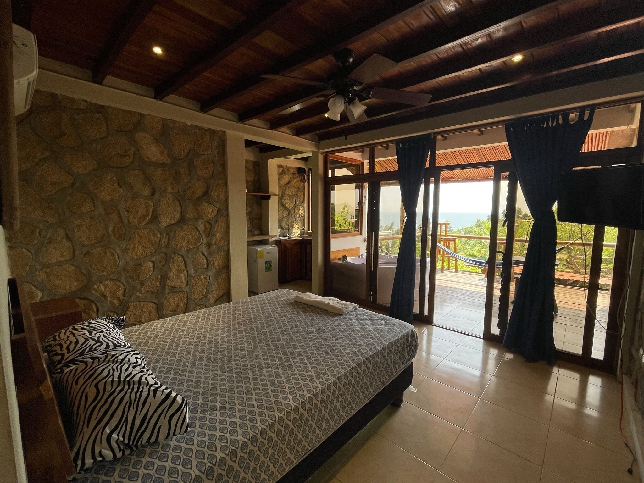 A bedroom with a bed, a stone wall, a ceiling fan, and a view of the outdoors.