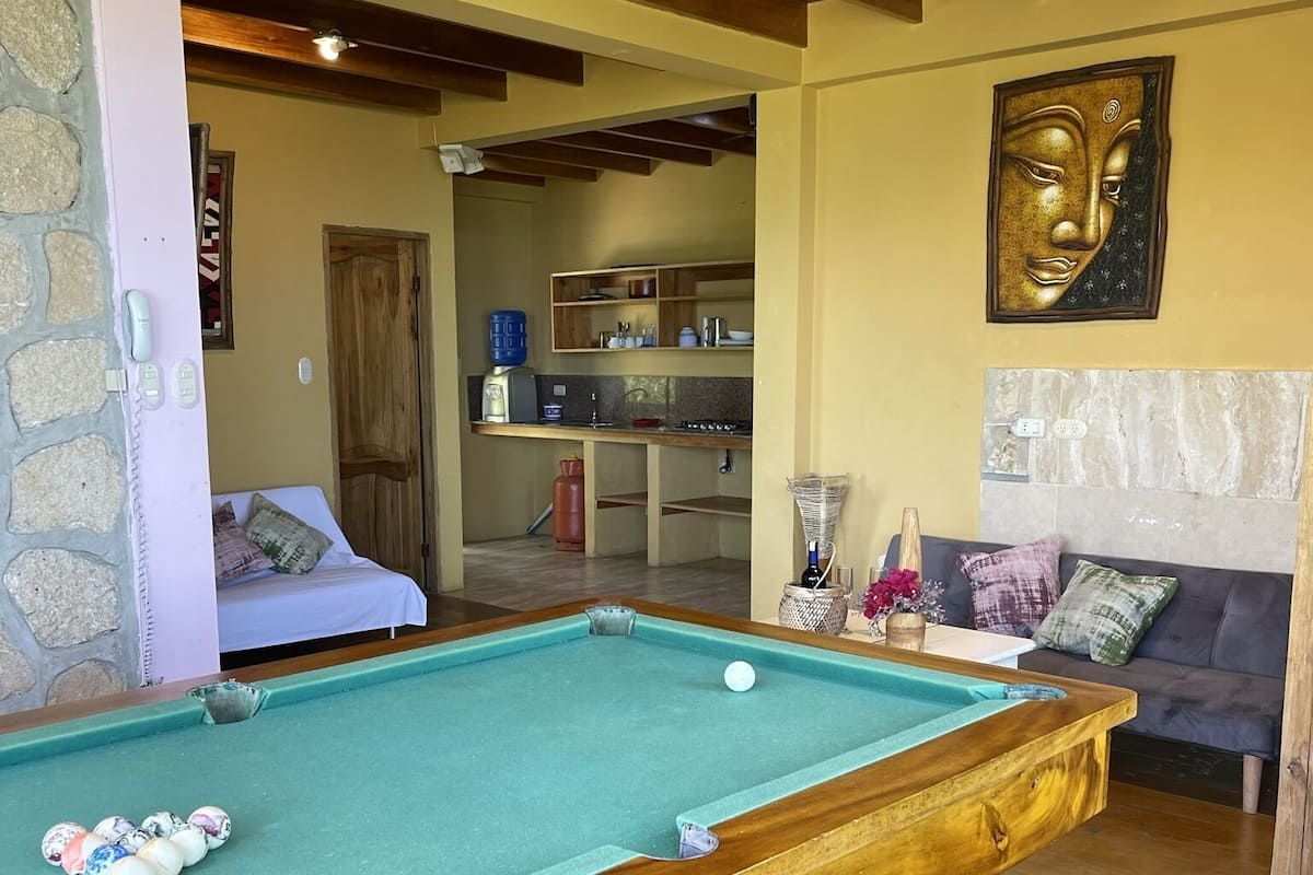 Une table de billard dans une pièce avec un mur en pierre, des poutres en bois au plafond, une statue de Bouddha et un canapé avec des coussins.