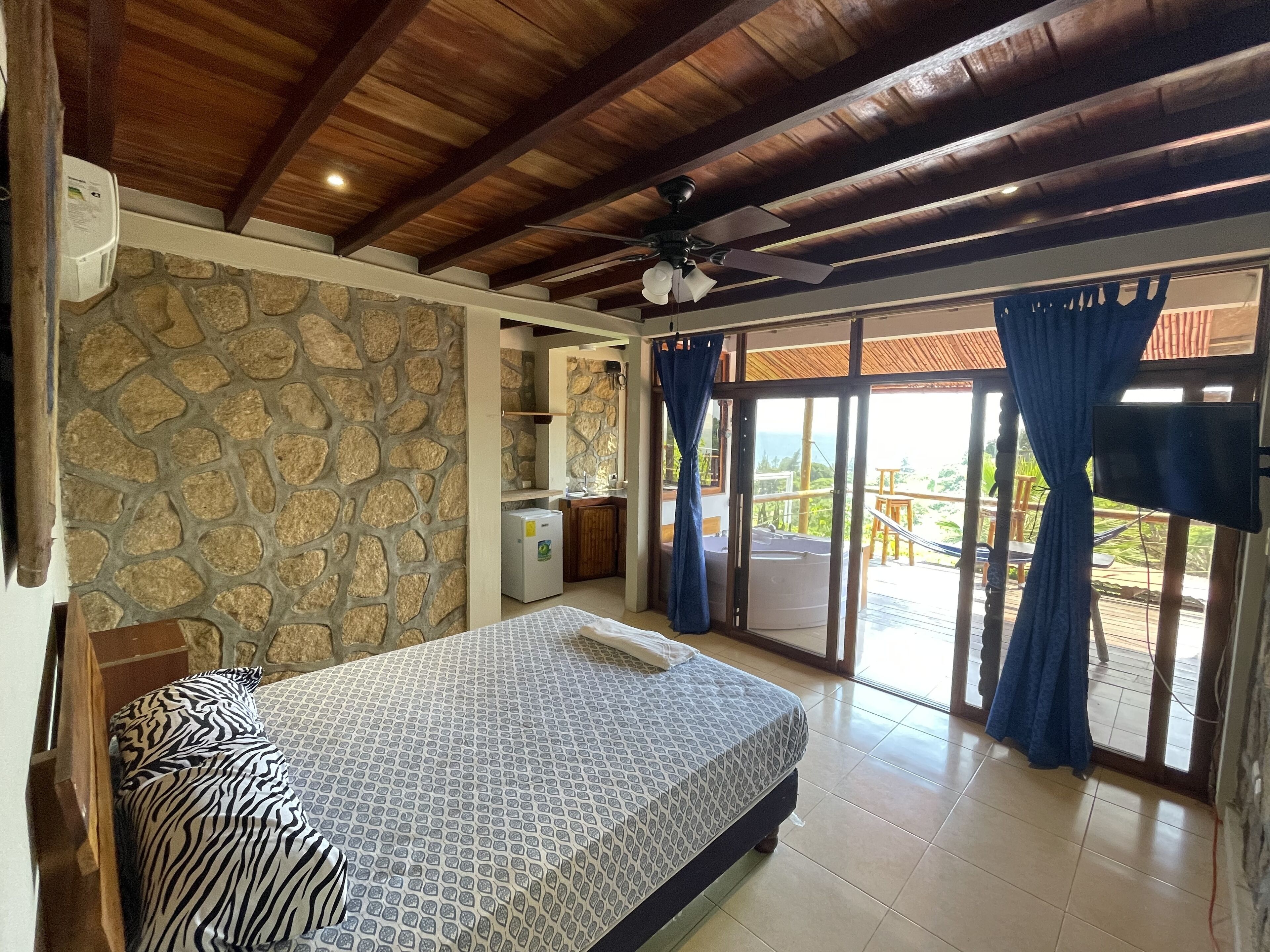 A bedroom with a stone wall, a bed with patterned bedding, a ceiling fan, and a view of the outdoors through a sliding door.