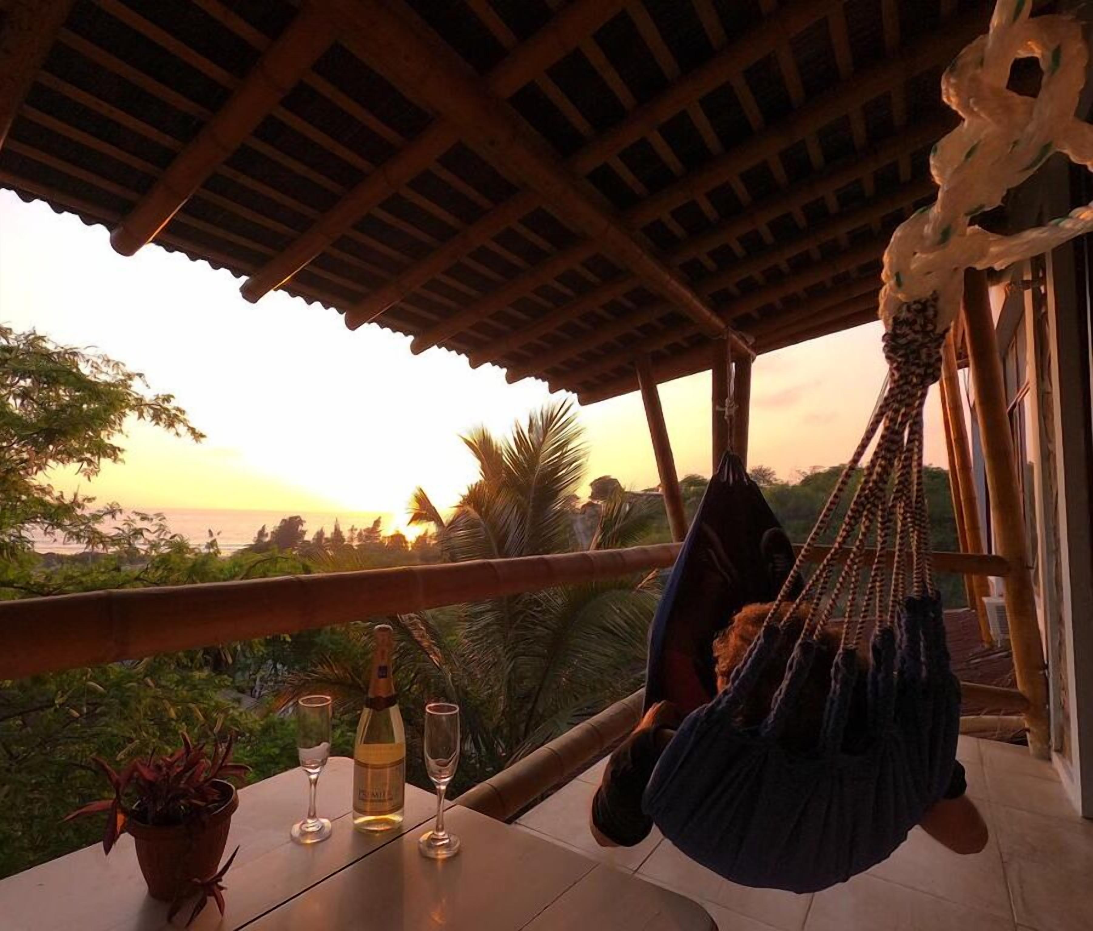 Una hamaca bajo una pérgola de madera con una mesa, copas de vino y una botella, con vistas a un paisaje exuberante al atardecer.