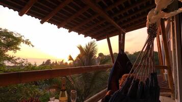 Una hamaca bajo una pérgola de madera con una mesa, copas de vino y una botella, con vistas a un paisaje exuberante al atardecer.